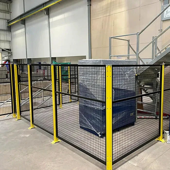 Black mesh fencing with yellow posts around a wrapped load beside factory stairs.
